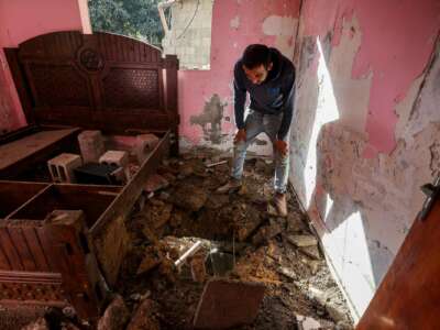 A man looks through a hole blown through his floor as he stands in a destroyed bedroom
