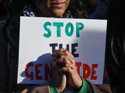 A person holds a sign reading "STOP THE GENOCIDE" behind their clasped hands