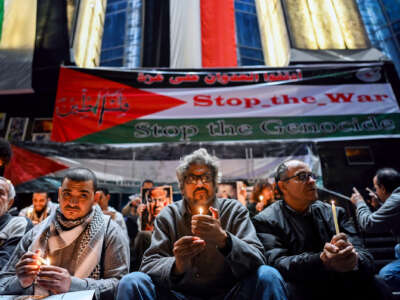 Men holding candles sit in front of a Palestinian flag bearing the words "STOP THE WAR; STOP THE GENOCIDE" during an outdoor vigil