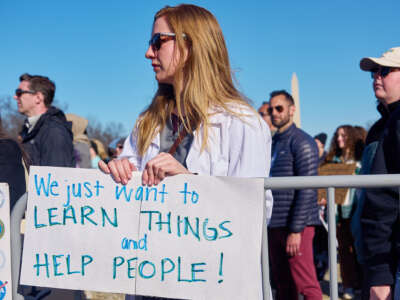 A scientist in her lab coat holds a sign reading "We just want to LEARN THINGS and HELP PEOPLE!" during an outdoor demonstration