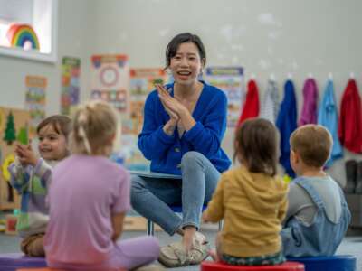 A preschool teacher sits in front of her students on the floor of the classroom as she prepares to read them a story. The children are each sitting on a circular cushion and are listening attentively to her instructions.