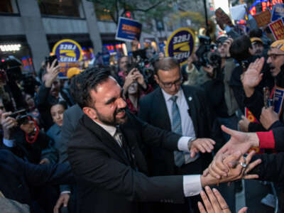 Zohran Mamdani is seen shaking hands with people as other people rally behind him with signs.